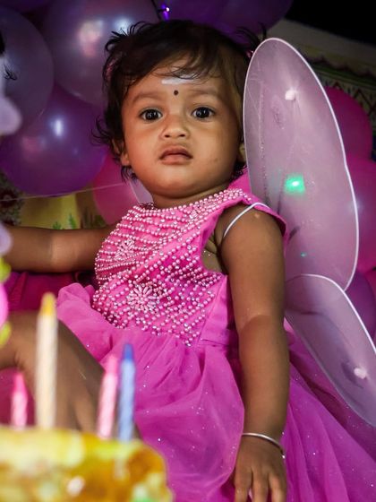 A magical moment from a first birthday party. The birthday girl, dressed in a pink fairy costume with little wings, looks on as the candles on her cake are lit.