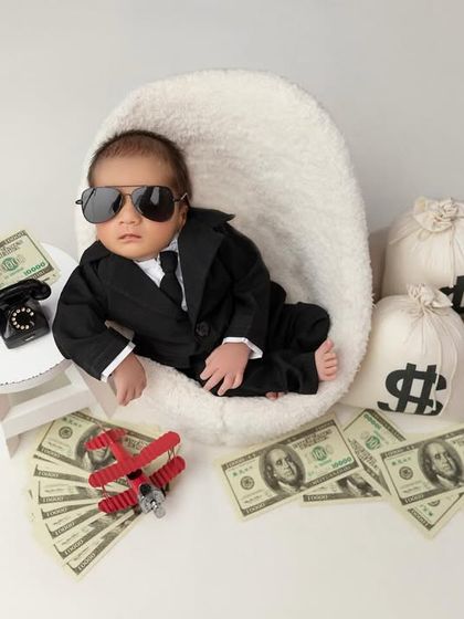 The boss baby has arrived. This fun and creative setup features a baby in a tiny suit and sunglasses, surrounded by prop money and bags, looking cool and in charge.