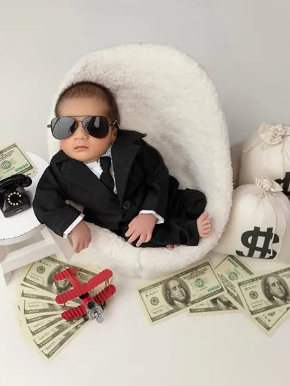 The boss baby has arrived. This fun and creative setup features a baby in a tiny suit and sunglasses, surrounded by prop money and bags, looking cool and in charge.