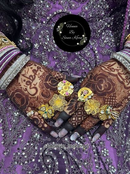 A stunning, dark henna stain on a bride's hands, adorned with beautiful gold rings. The Arabic calligraphy adds a personal and elegant touch.