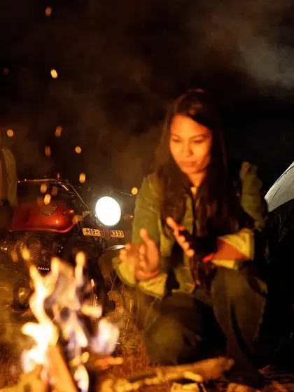 A candid moment by the fire. She warms her hands by the flames while he watches from beside their motorcycle, the scene lit by the campfire and the bike's headlamp.