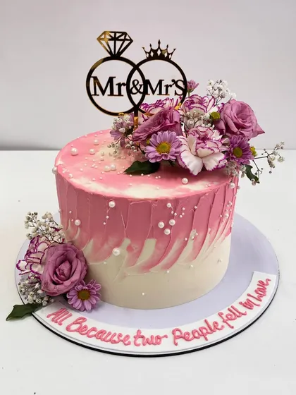 "All Because two People fell in love." This romantic engagement cake features a soft pink and white watercolor effect, delicate florals, and a sweet message written on the board.