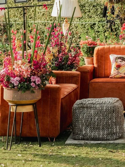 A comfortable and stylish lounge corner with a plush orange sofa, a knitted pouf, and a tall floral arrangement. The perfect spot for guests to relax and chat.