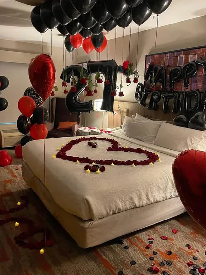 A dramatic red and black themed 27th birthday decoration in a hotel room, featuring a rose petal heart and number balloons with hanging roses.