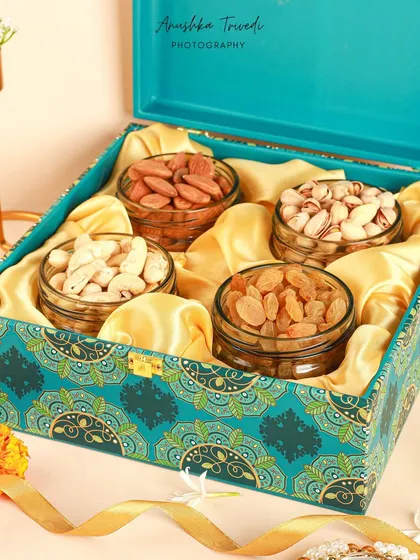 An open dry fruit hamper for Diwali, showing the goodies inside. The turquoise box with its intricate pattern is complemented by the warm tones of the nuts and diyas.