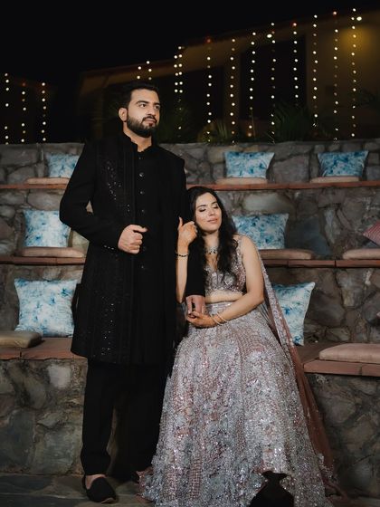 A classic, posed portrait of the couple at their sangeet. Their elegant black and silver outfits stand out against the rustic stone and warm fairy lights.