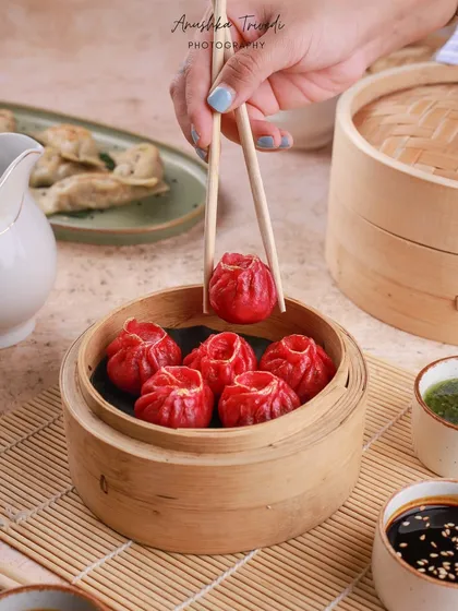 Vibrant red dumplings in a bamboo steamer. The hand holding one with chopsticks adds a human element and shows the delicate shape of the dumpling.