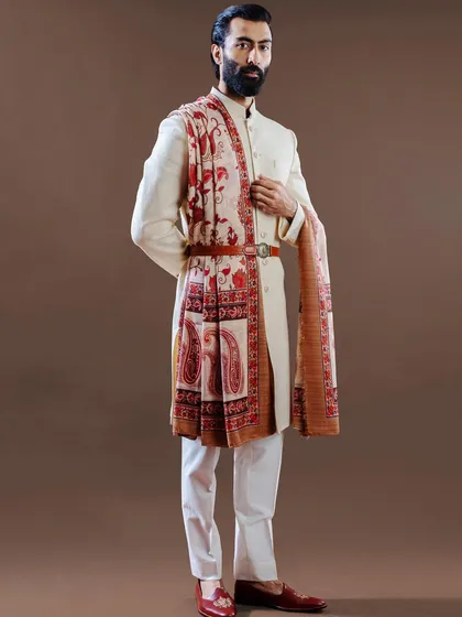 A bold statement in wedding fashion, this ivory achkan is paired with a richly printed pashmina shawl, secured by a leather belt. This look masterfully blends traditional textiles with a contemporary styling sensibility.