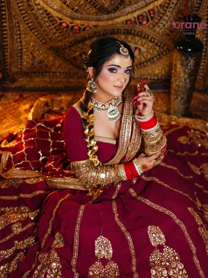 A classic seated bridal pose, showcasing the rich maroon lehenga, intricate gold embroidery, and flawless makeup.