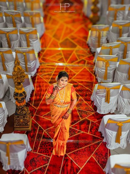 A high-angle shot of the bride, Dhanya, standing in the aisle of the wedding hall. This perspective emphasizes the vibrant geometric patterns of the carpet and the elegant decor, creating a powerful solo portrait.