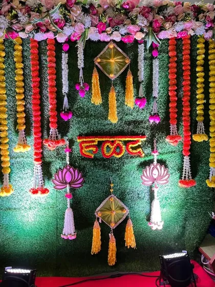 A traditional Haldi backdrop made of a lush green wall. It's decorated with marigold and rose flower strings, along with decorative hangings.