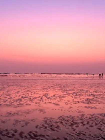 A beautiful photograph of a Mumbai beach at sunset, with the wet sand reflecting the soft pink and purple hues of the sky.