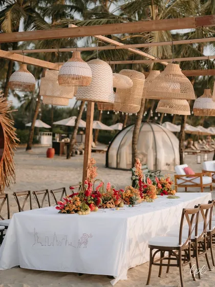 A long dining table set up for a beach party, with hanging rattan lamps and a custom-illustrated table runner.
