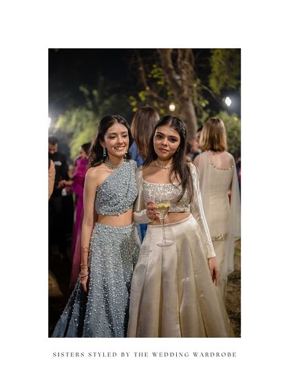 Sisters styled by us for a sangeet night. One wears a chic one-shoulder lehenga in dusty blue, while the other is in a classic ivory lehenga, showcasing how individual styles can still look cohesive.