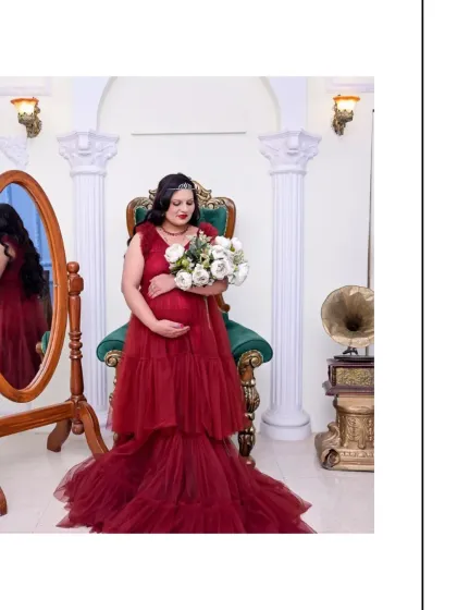 A serene moment in our royal-themed setup. The mom-to-be, dressed in a rich red gown, holds a bouquet of white flowers while seated on the throne chair, with her reflection captured in the vintage mirror.