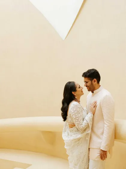 A sweet, intimate moment between the couple in their elegant white outfits. The simple, neutral background puts all the focus on their loving gaze and gentle interaction.