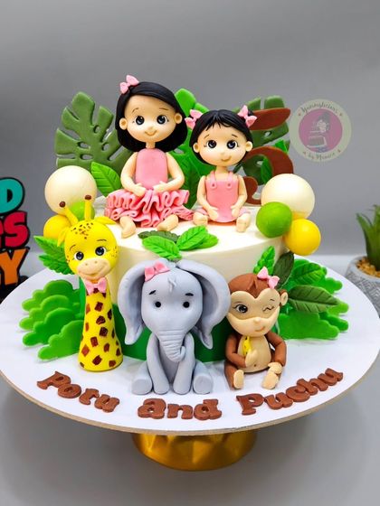 A jungle-themed cake for two sisters, "Peru and Puchu," featuring custom figurines of the girls alongside cute fondant animals.