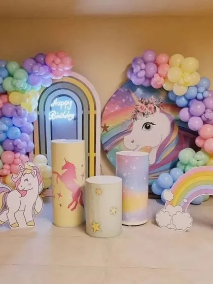 A magical unicorn and rainbow themed first birthday. The setup includes pastel balloon arches, unicorn cutouts, a rainbow prop, and themed pedestals.
