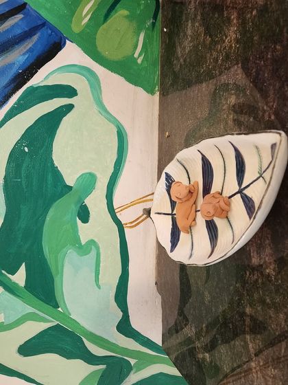 A painted leaf tray with two small clay figures, set against a vibrant mural, blending guest art with our cafe's decor.