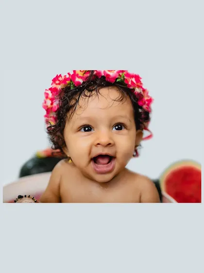 A close-up shot of a baby girl's expressive face, crowned with a floral headband during her watermelon-themed photoshoot. Her wide eyes and happy smile are simply captivating.