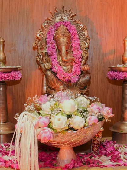 A close up of a Ganpati idol adorned with a fresh pink flower garland. The setup includes traditional brass lamps (samai) and a bowl of lotuses, creating a simple yet beautiful space for worship.