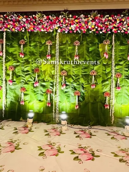 A classic South Indian backdrop made from fresh banana leaves. The stage is decorated with hanging floral tassels and a top border of colorful flowers, perfect for any auspicious event.