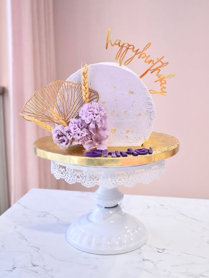 A dreamy lilac-colored cake with modern, artistic decorations. This design features a textured disc, dried wheat stalks, and delicate purple flowers, all accented with a gold fan.