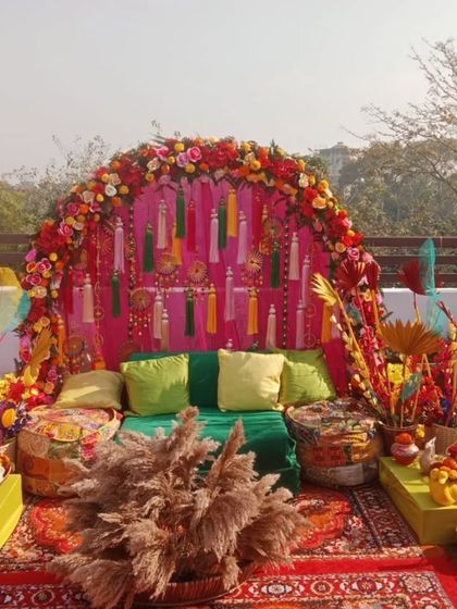 A vibrant and festive rooftop Mehandi setup with low gadda seating and colorful cushions. The backdrop is a floral arch with tassels, surrounded by baskets of flowers and pampas grass.