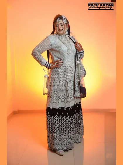 A full-length portrait of the bride, showcasing her magnificent wedding lehenga. The warm, orange-toned background makes her silver and blue outfit shine.