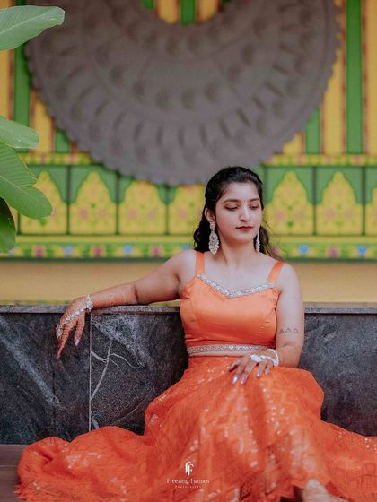 A relaxed portrait of the bride in her orange Haldi dress, seated against a colorful, traditional backdrop.