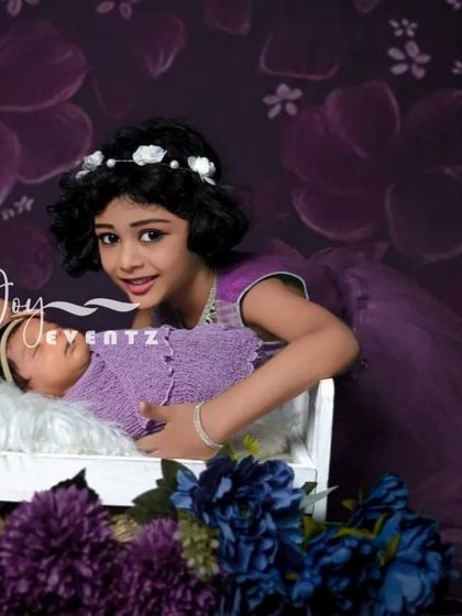 A lovely sibling photoshoot with a violet theme. The older sister looks lovingly at her newborn sibling, who is resting in a miniature cot, all set against a beautiful floral backdrop.