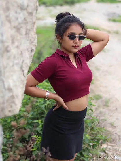 A cool and casual outdoor shot of a model in a maroon crop top, black skirt, and sunglasses. The pose is relaxed and confident.