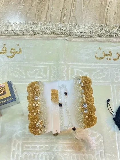 A personalized prayer set featuring a white and gold resin Quran holder with a crystal tasbeeh. The soft, light-colored prayer mat is customized with names in glittering gold script.
