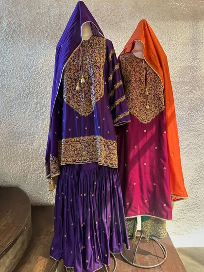 Two regal sharara sets ready for a grand celebration. The deep purple and magenta outfits feature heavy gold zardozi work on the neckline and sleeves, paired with flowing dupattas.