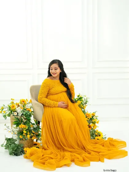 A bright and sunny portrait of an expecting mother in a beautiful yellow lace gown. She is seated and surrounded by baskets of yellow and white flowers, creating a cheerful and fresh look.