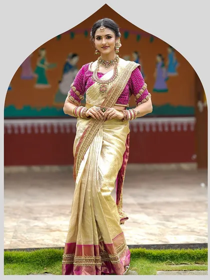 A full-length view of a classic South Indian bridal look. The bride wears a cream and gold silk saree with a richly embroidered magenta blouse, standing against a backdrop that evokes traditional charm.
