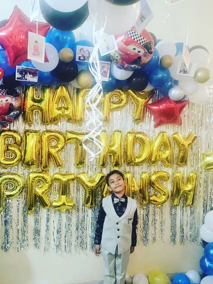A 'Cars' themed 7th birthday party. The birthday boy stands proudly in front of a silver fringe backdrop, framed by a balloon arch with character cutouts and photos.