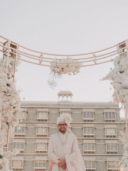 The groom awaits his bride, standing under a magnificent floral arch that perfectly frames the grandeur of the palace hotel.