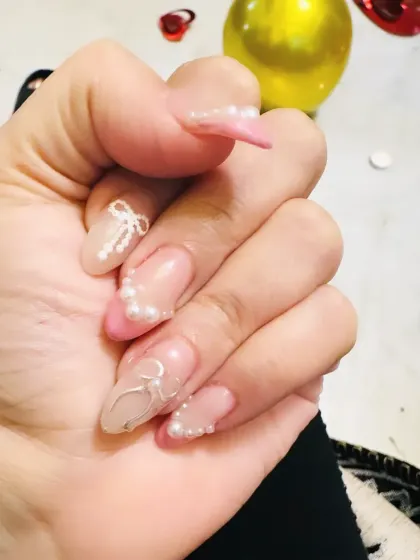 A delicate and romantic design on almond-shaped nails, featuring hand-painted white bows and scattered pearls on a sheer pink base.