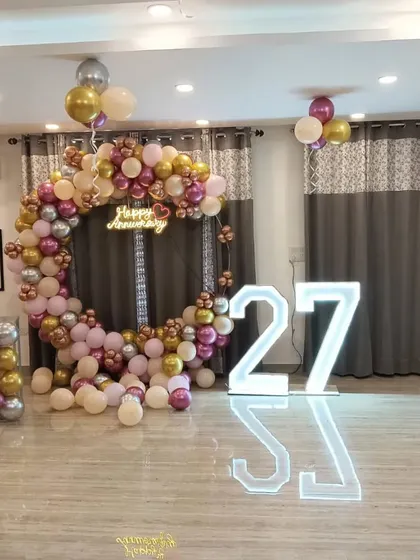 A 27th-anniversary celebration with a circular balloon arch in pink, gold, and silver. The setup includes large marquee numbers and floating balloons.