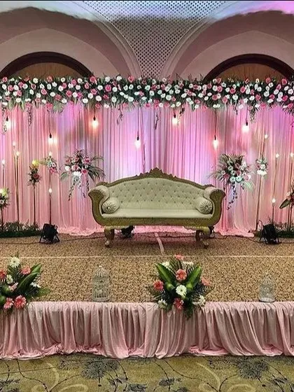 Here is a reception stage setup with a soft, romantic feel. I used a backdrop of pink drapes with hanging bulb lights, topped with a dense arrangement of pink and white flowers to create a warm and inviting space.