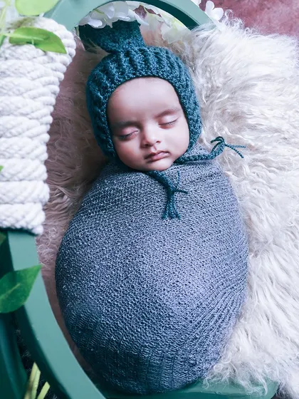 A close-up shot of a baby swaddled in a grey wrap and bonnet, sleeping soundly in a basket. The textures of the knitwear and fur create a cozy feel.