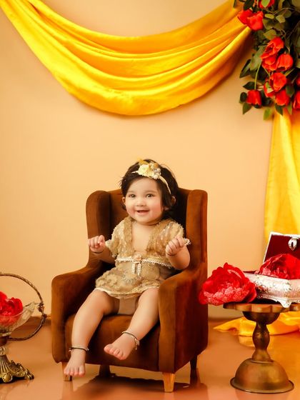 A smiling baby girl sits in a miniature armchair, surrounded by a golden backdrop and red roses. This setup is perfect for a grand, royal-themed shoot.
