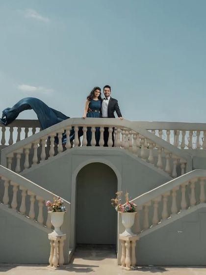 A wide shot of a couple on a grand staircase set, showcasing the scale of the location and the elegance of their attire.