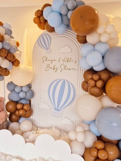 A dreamy hot air balloon themed baby shower with a teddy bear pilot. The setup uses a soft color palette of blue, brown, and white balloons to create a sky-like atmosphere with fluffy clouds at the base.