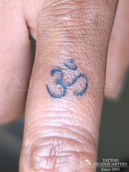 A simple and clean Om symbol tattooed on the thumb, a popular placement for a personal and constant spiritual reminder.