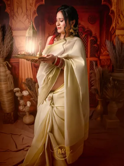 A serene and spiritual-themed maternity portrait. The mother-to-be, dressed in a traditional white saree, holds a lit diya, creating a peaceful and culturally significant image.