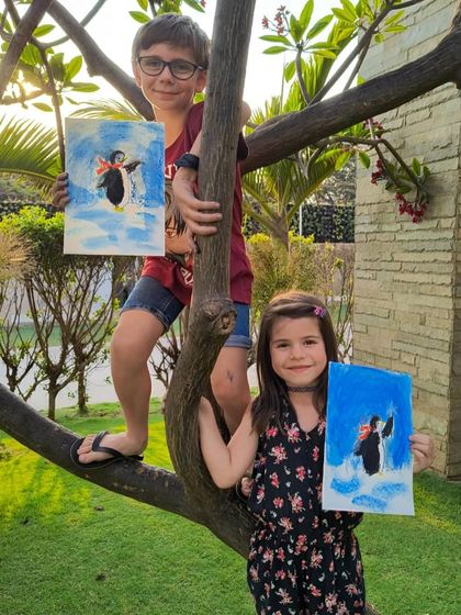 Two students climb a tree to show off their penguin paintings, mixing art with a little adventure.