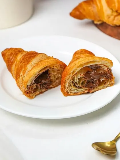 A Nutella-filled croissant cut in half, with a second croissant and a coffee pot in the background. This shot creates a lovely breakfast or coffee break scene.