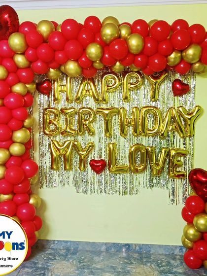 A simple and classic "Happy Birthday My Love" home decor setup. The red and gold balloon arch frames a silver tinsel curtain, perfect for an intimate celebration.